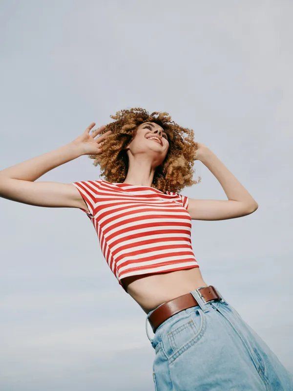 A young woman in a red and white striped crop top and denim jeans poses outdoors against a clear blue sky, radiating carefree energy and confident summer style.