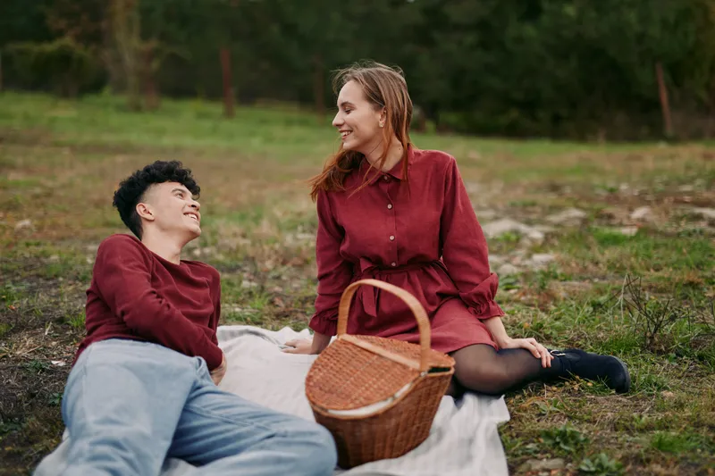 Candid outdoor couple picnic on a blanket with a wicker basket, showing genuine smiles and natural interaction that conveys authenticity and credibility in warm, everyday romance moments.