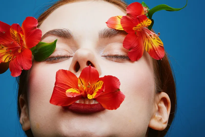 Close up portrait of a model with flowers covering her lips, vibrant colors, expressing beauty and femininity, with a blue background, perfect for concepts of wellness and nature