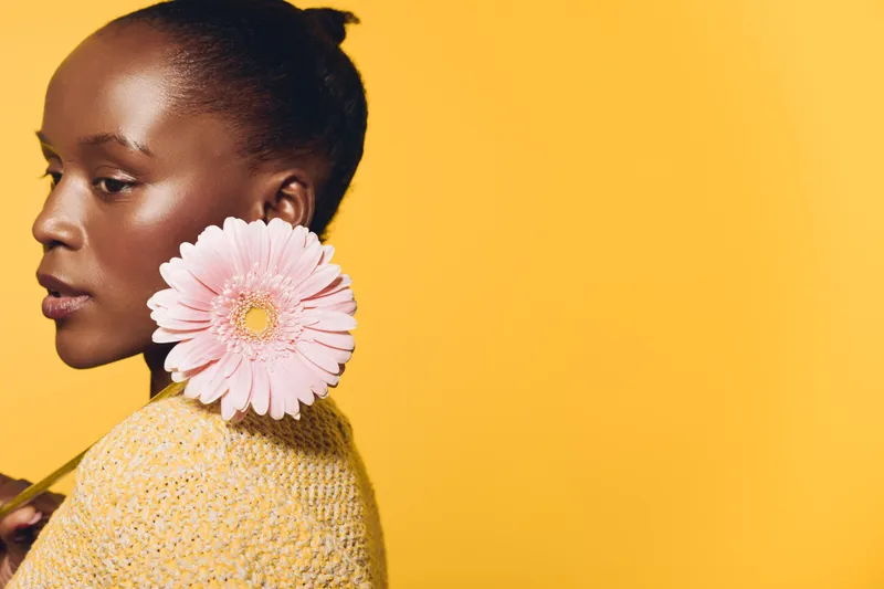 Portrait of a young black woman with dark skin holding a large pink flower near her face against a bright yellow background. Concept of beauty, natural elegance, and floral aesthetics.
