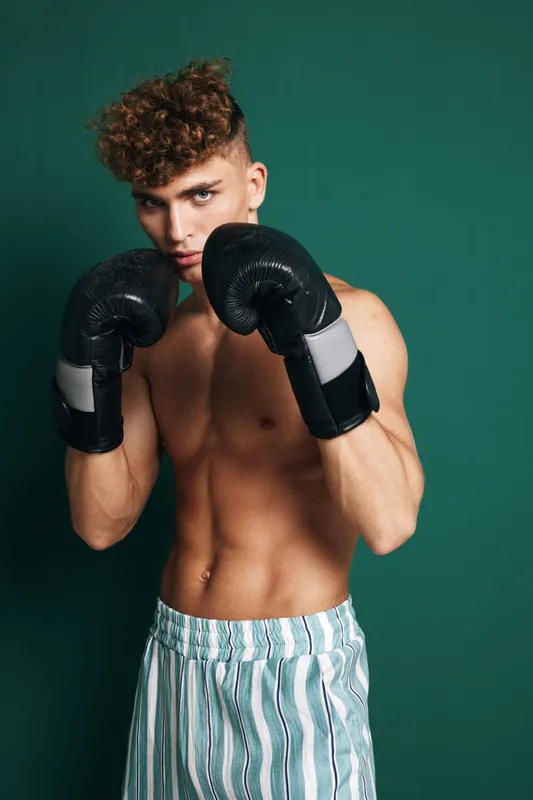 Young male boxer with curly hair wearing black gloves and striped shorts posing in fighting stance against green background for fitness and training concept.