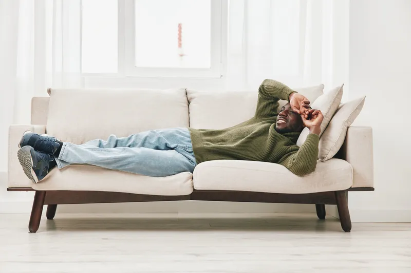 Relaxed Black man lying on a cozy sofa, wearing a green sweater and jeans, looking contemplative in a bright living room