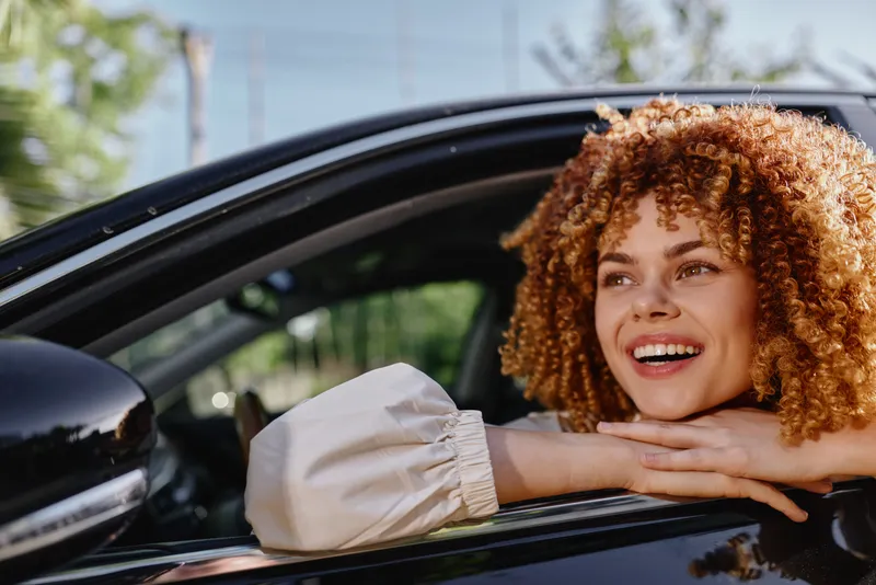 Smiling woman with curly hair leaning out of car window enjoying sunny day, wearing light blouse, relaxed and happy in natural outdoor setting.