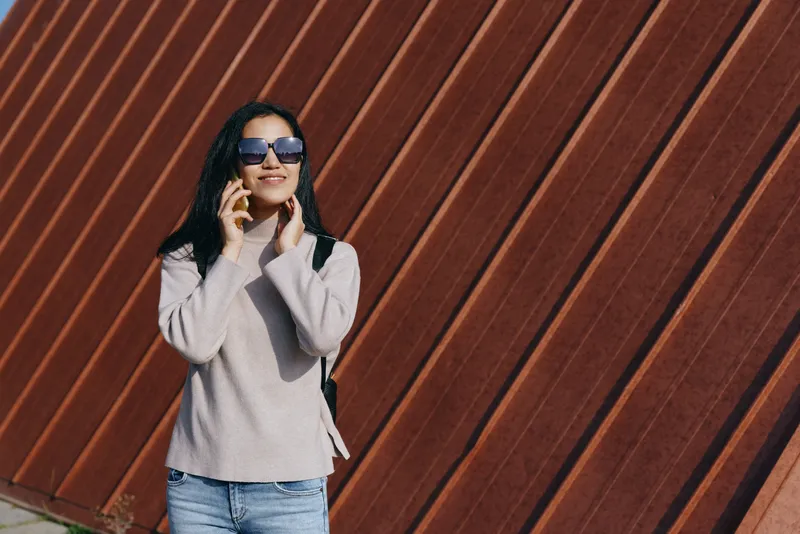 Woman wearing sunglasses talks on mobile in casual outfit beside a diagonal timber wall, showcasing street style, outdoor vibe and modern fashion with confident everyday communication