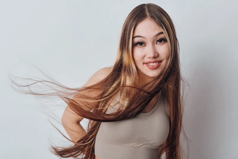 Young woman with long hair smiling and posing in casual tank top against white background. Female model with natural makeup and flowing hair in studio.