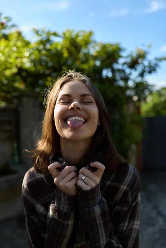 Playful woman outdoors smiles with tongue out, wearing plaid shirt, close up portrait, natural light, casual lifestyle moment in sunny backyard garden. outdoors
