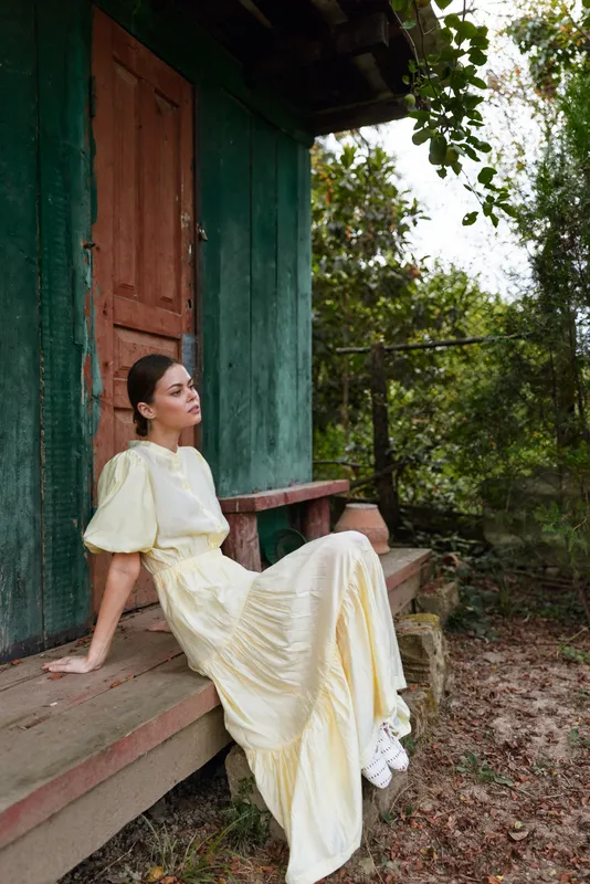 Woman sits gracefully on a rustic porch wearing a pale yellow dress, relaxed pose beside a green dwelling, capturing a serene outdoor portrait with soft natural light for a timeless vibe and warm