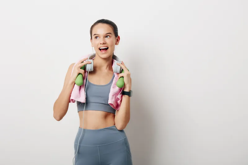 Fitness woman in athletic wear with a towel around her neck, lifting small green dumbbells and smiling, energized during a studio workout in a clean, minimal space.