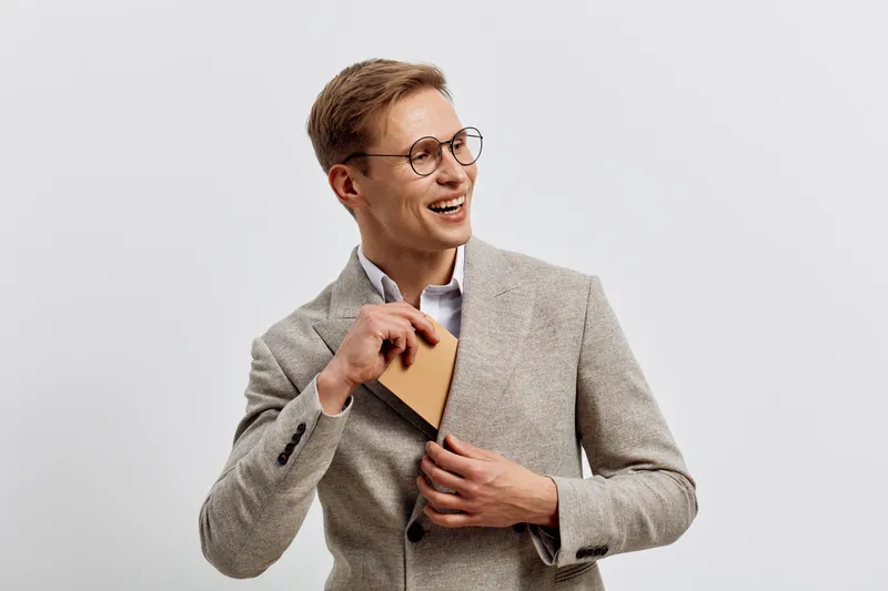 Smiling young Caucasian man wearing glasses and a gray blazer holding a notebook, looking happy and confident, isolated on plain background. People lifestyle concept, professional, cheerful, modern st