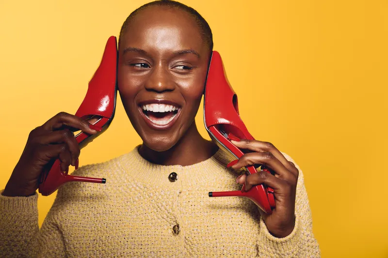 Happy black woman with dark skin holds red high heels near ears smiling broadly in yellow studio. Concept of joy, fashion, beauty, and playful portrait of African ethnicity female model.