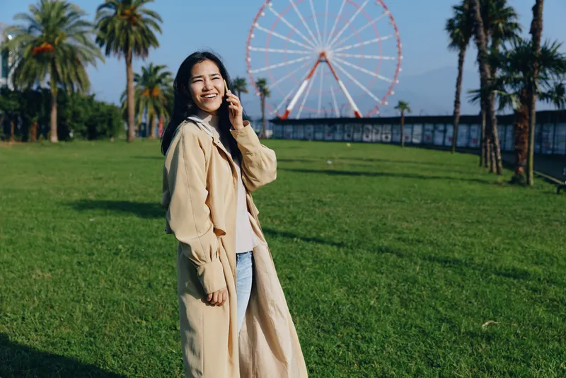 Woman in beige coat walking in a sunny park with a ferris wheel in the background, casual outdoor lifestyle, bright daylight, smiling expression and relaxed mood.