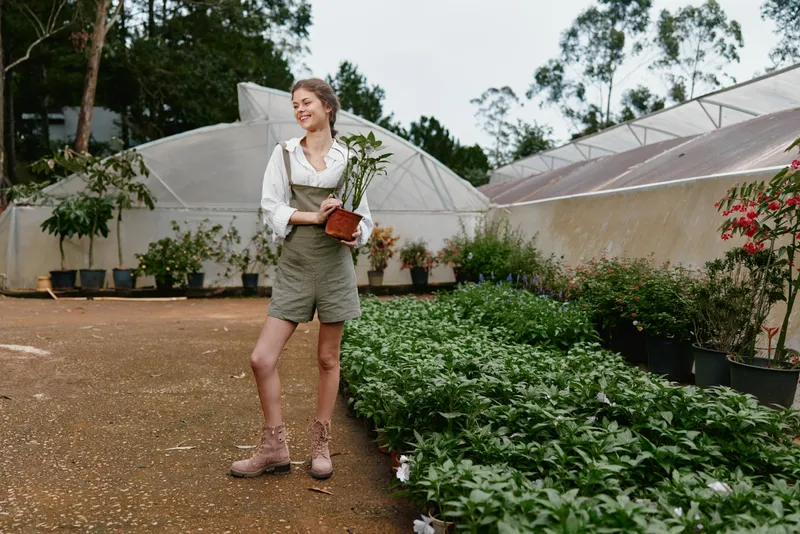 Young female gardener in casual outfit smiling with potted plant in greenhouse, exuding joy in nature, representing sustainable gardening and plant care