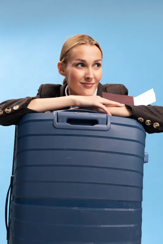 Young woman leaning on large blue suitcase with travel documents in hand, smiling and looking away. Concept of travel preparation, vacation, and anticipation of journey.