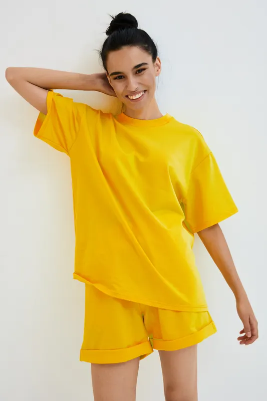 smiling woman in yellow casual outfit posing against white wall, short hair tied up, cheerful expression, relaxed mood, indoor minimalistic background