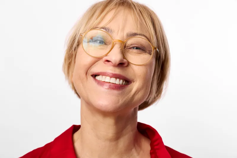 Woman with glasses smile in a closeup portrait, blonde short hair, red clothing, eyewear, friendly expression, studio shot on clean white background, soft light