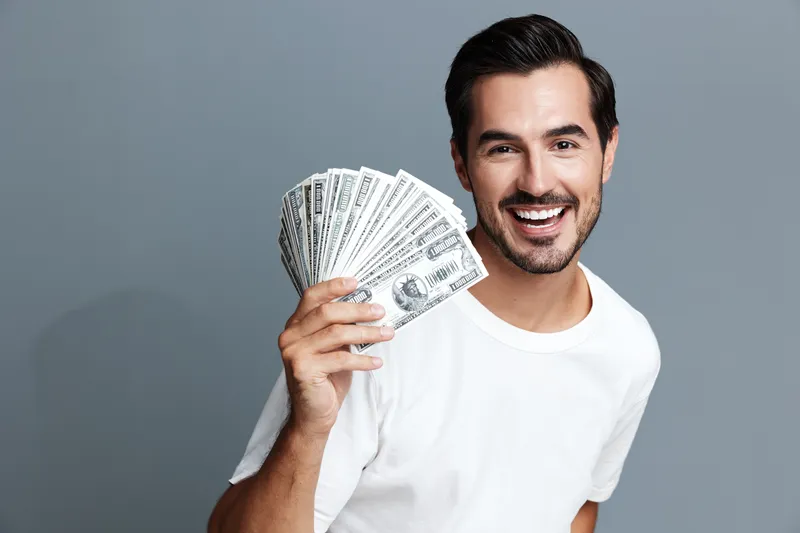 Happy smiling man holding cash in hand, showcasing excitement and joy, dressed in a casual white t-shirt, against a solid gray background. Financial success concept.