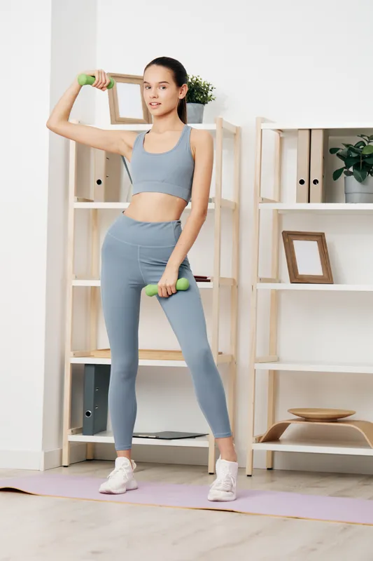 Woman fitness athlete lifting light dumbbells in a bright home gym, wearing blue activewear, showcasing a confident pose with shelves and decor in the background.