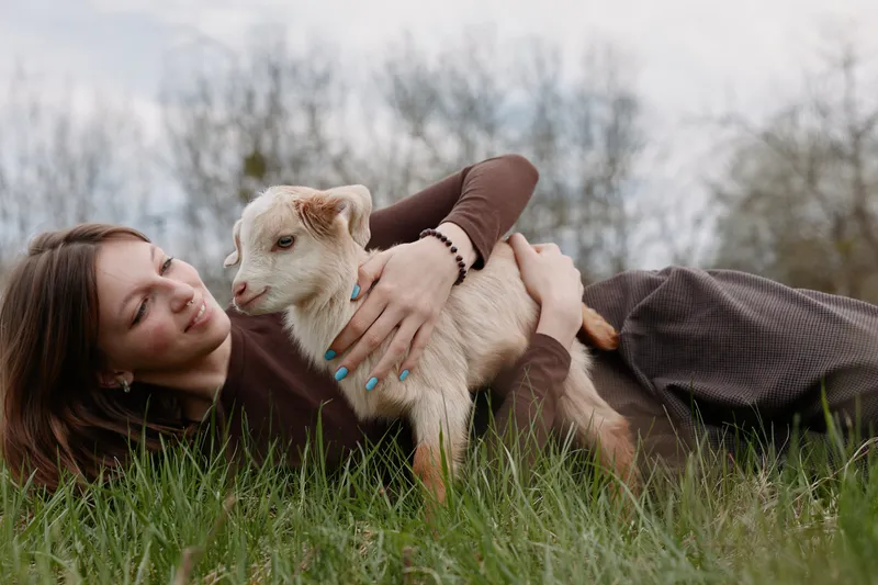 Joyful woman outdoors, lying on grass, embracing a curious goat kid, gentle interaction, soft natural lighting, peaceful countryside vibe, candid lifestyle moment, warmth and connection