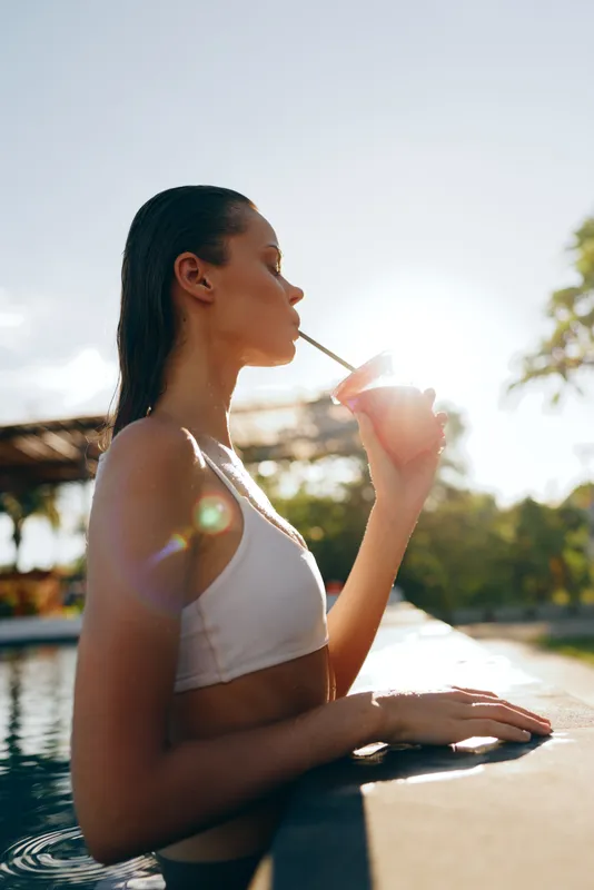 Relaxed woman leans on pool edge, sipping through a straw, soaking up sun by a bright pool, wearing a white bikini, candid lifestyle moment, serene outdoor setting.