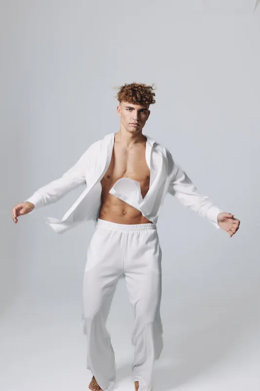 Young man with curly hair wearing white open shirt and pants posing in studio with neutral background for fashion, style and casual outfit concept.
