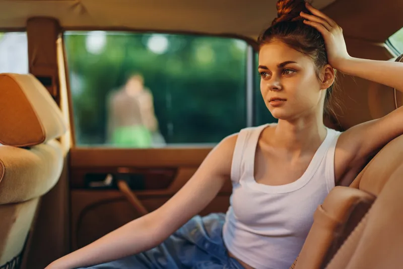 Young woman in casual outfit sitting in the backseat of a car, looking contemplative and relaxed, while soft natural light illuminates the scene.
