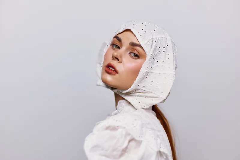 A portrait of a young woman with a vintage white bonnet, showcasing a soft expression against a neutral grey background, ideal for fashion and cultural themes