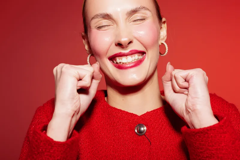 Happy woman in red sweater smiles with closed eyes and clenched fists on red background. Concept of joy, excitement, celebration, and positive emotions in studio lighting.