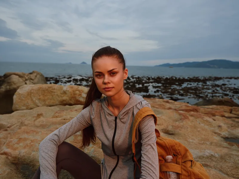 Young woman in sporty outfit sitting on rocky beach, showcasing determination against the ocean backdrop at sunset, capturing the spirit of adventure and exploration