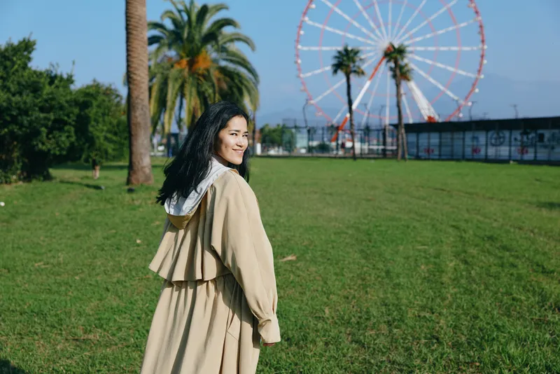 Woman in a beige coat walking through a sunny park with green grass, palm trees, and a distant ferris wheel, capturing a calm, casual moment in an outdoor setting