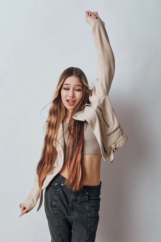 Young woman with long hair wearing casual jacket and jeans dancing with raised arm and closed eyes isolated on white background in motion for lifestyle and fashion concept.