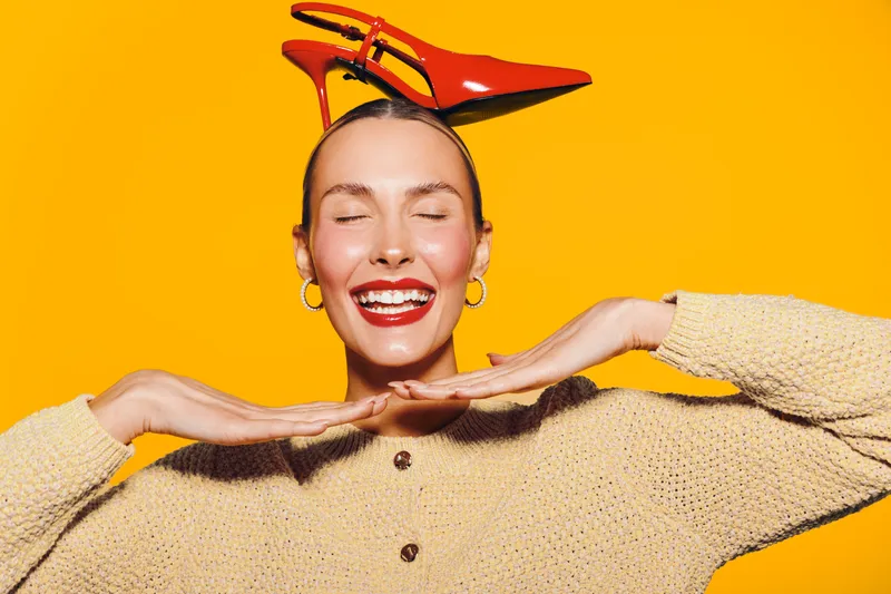Happy woman with red lipstick and earrings smiles with closed eyes, posing with red high heel shoes on her head against bright yellow background. Concept of joy, fashion, and fun.