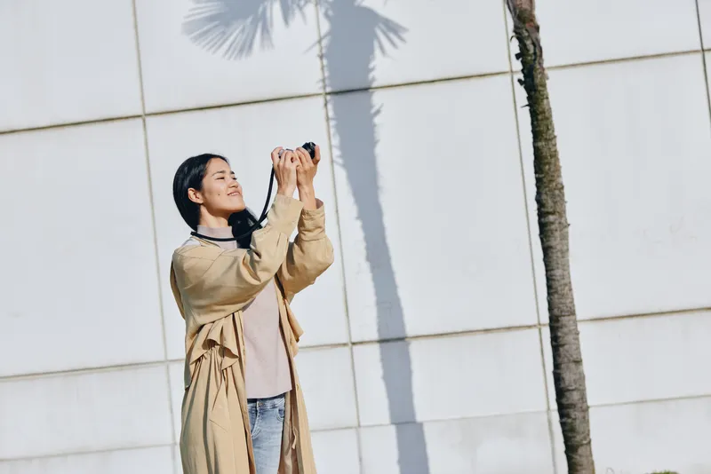 Woman with camera shoots upward outdoors beside a palm tree and a clean wall, capturing a candid moment in daylight, showcasing photography, travel, and everyday creativity
