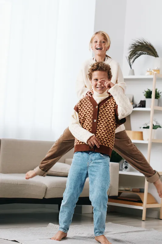Playful siblings share a joyful indoor moment as a girl sits on a boy's shoulders, both smiling and leaping in a bright, cozy living room.