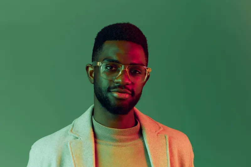 Confident young African man with glasses wearing stylish casual clothes against a green background, exuding positivity and sophistication