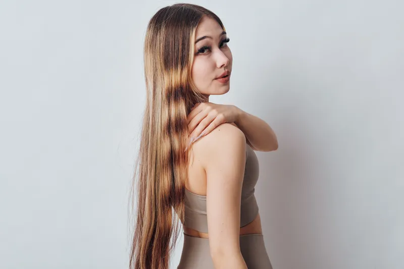 Young woman with long straight hair and neutral makeup posing in beige sporty outfit on plain background. Female model showing hairstyle and smooth skin, beauty and fashion concept.