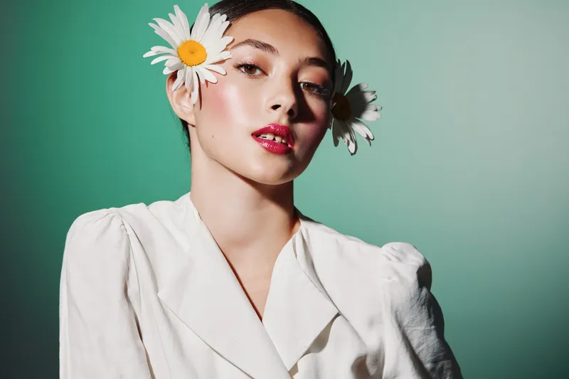 Fashionable young woman with pink lipstick and flowers in hair poses looking at camera against a green background. Concept for beauty creative and fashion aesthetic.