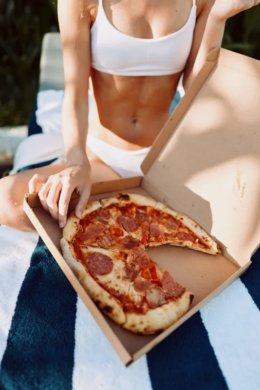 Outdoor scene with a woman in a white bikini top enjoying pizza from an open box on a striped blanket, sunlit moment, relaxed posture, casual lifestyle, vibrant summer colors.