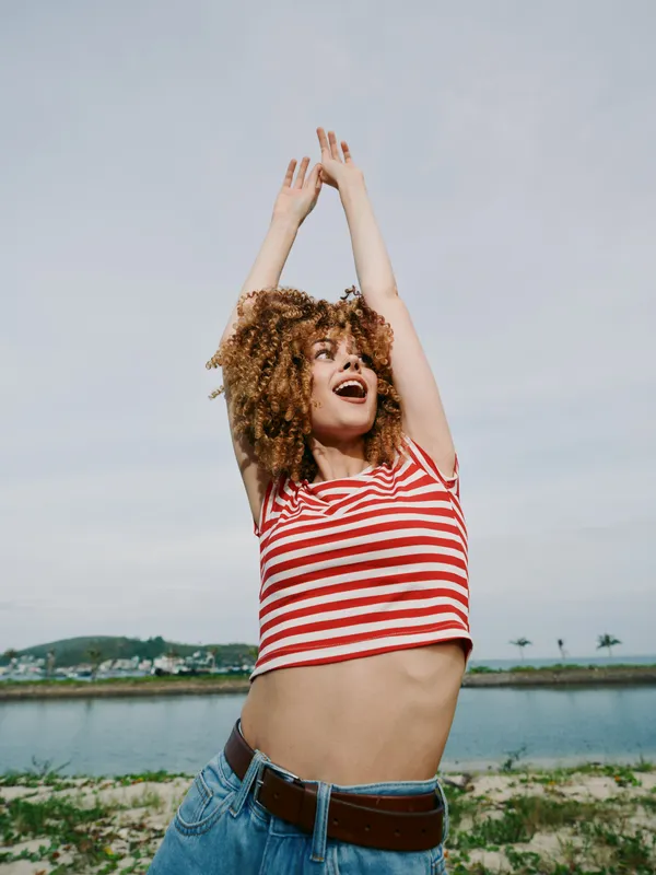 Playful young woman in a red and white striped shirt dances with raised arms by a lake, expressing joy, freedom, and carefree summer energy under a bright blue sky.
