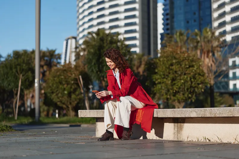 Young woman with curly hair sits on a concrete bench in an urban park, wearing a red coat and white pants, checking her phone with a focused smile.