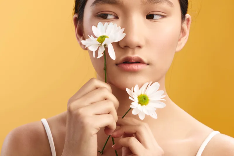 asian woman gracefully holds white daisies near her face against a warm orange backdrop, creating a soft, contemplative portrait that emphasizes natural beauty and calm emotion.
