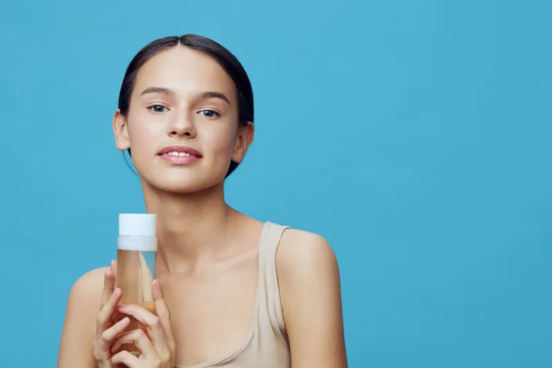 Young woman promoting natural skincare product with a friendly smile, showcasing beauty amidst a soft blue background and simple attire, ideal for wellness theme