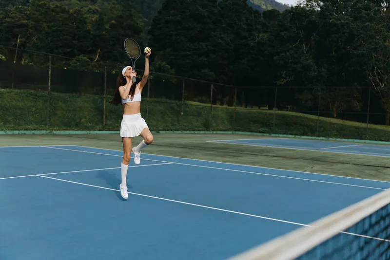 Young woman playing tennis in a stylish white outfit, showcasing excitement and concentration on a blue court surrounded by greenery, ideal for sports and fitness themes
