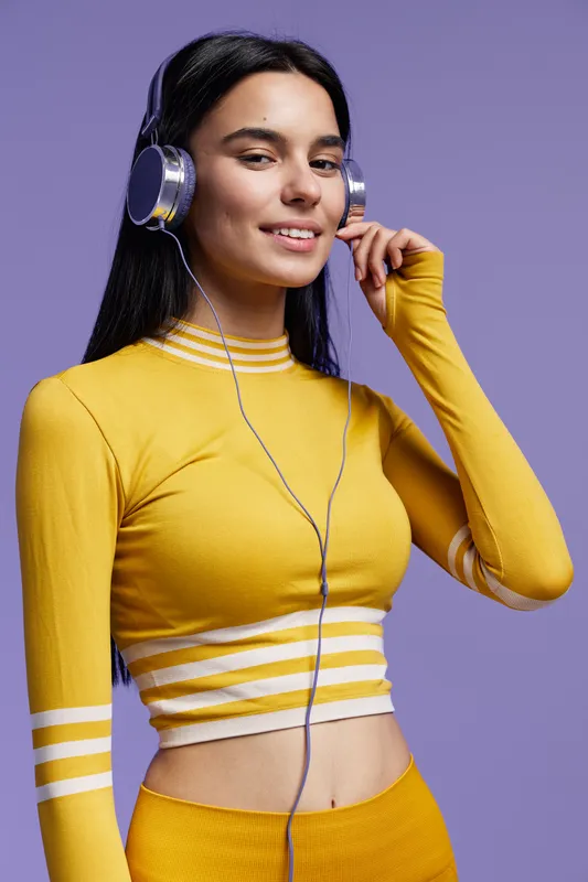 Young woman sport fitness wearing yellow activewear and headphones, posing confidently with a relaxed smile against a solid purple background.