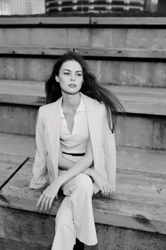 Black and white portrait of elegant woman sitting on wooden stairs wearing light suit and top, contemplative expression, urban fashion style