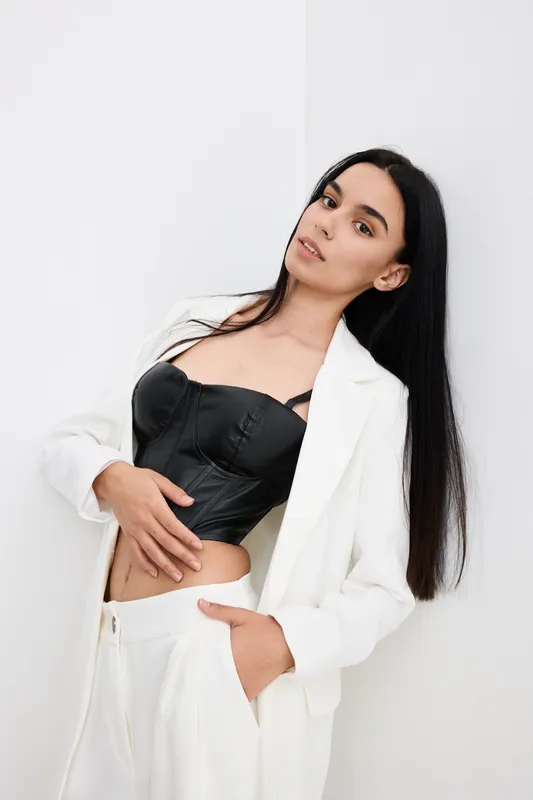 Stylish woman in white suit and black corset poses with calm expression against minimal white background, showcasing modern fashion and elegance with natural beauty.
