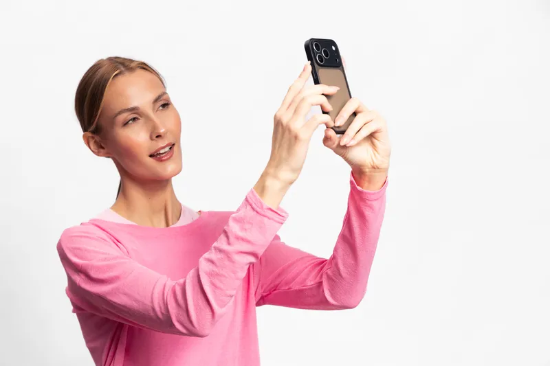 Young woman in pink sweater takes selfie with smartphone on white background. Casual lifestyle concept with natural makeup and relaxed expression in bright studio setting.