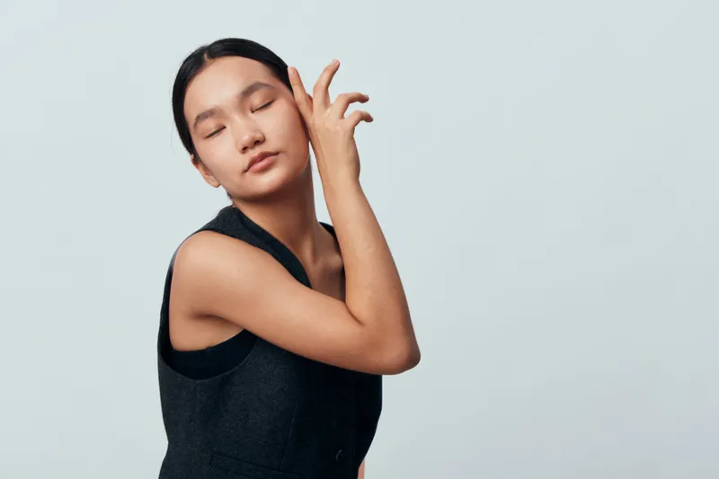 Young woman with closed eyes posing with hand near face in studio, wearing sleeveless dark top against plain light background with calm and peaceful expression.