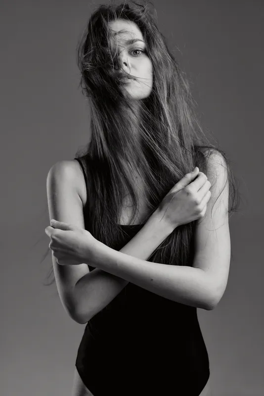 Black and white portrait of a young woman with long hair in a black outfit, expressing mystery and elegance with crossed arms.