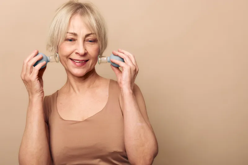 Smiling mature woman with short blonde hair using handheld facial massager on cheeks in neutral studio setting. Concept of skincare, anti aging, wellness, and self care for older adults.