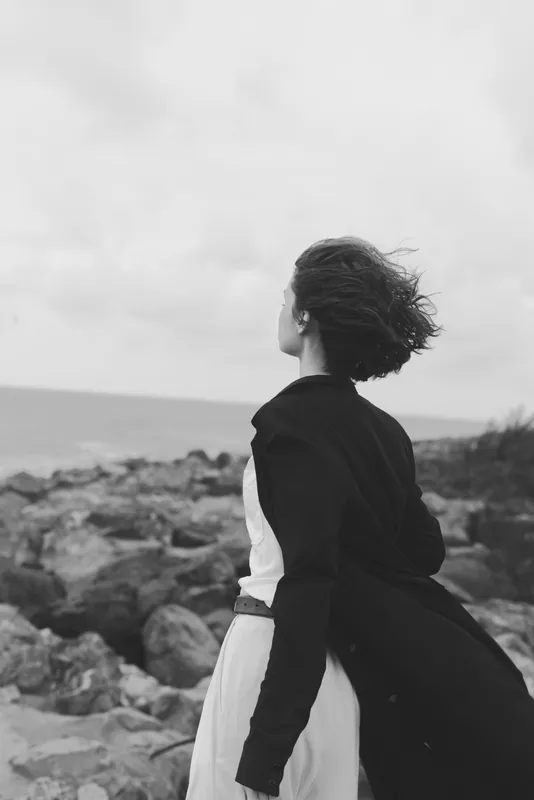 Windy coast scene with a woman standing in a flowing dress and jacket, looking toward the sea, rocky shoreline, monochrome mood, sense of solitude and freedom